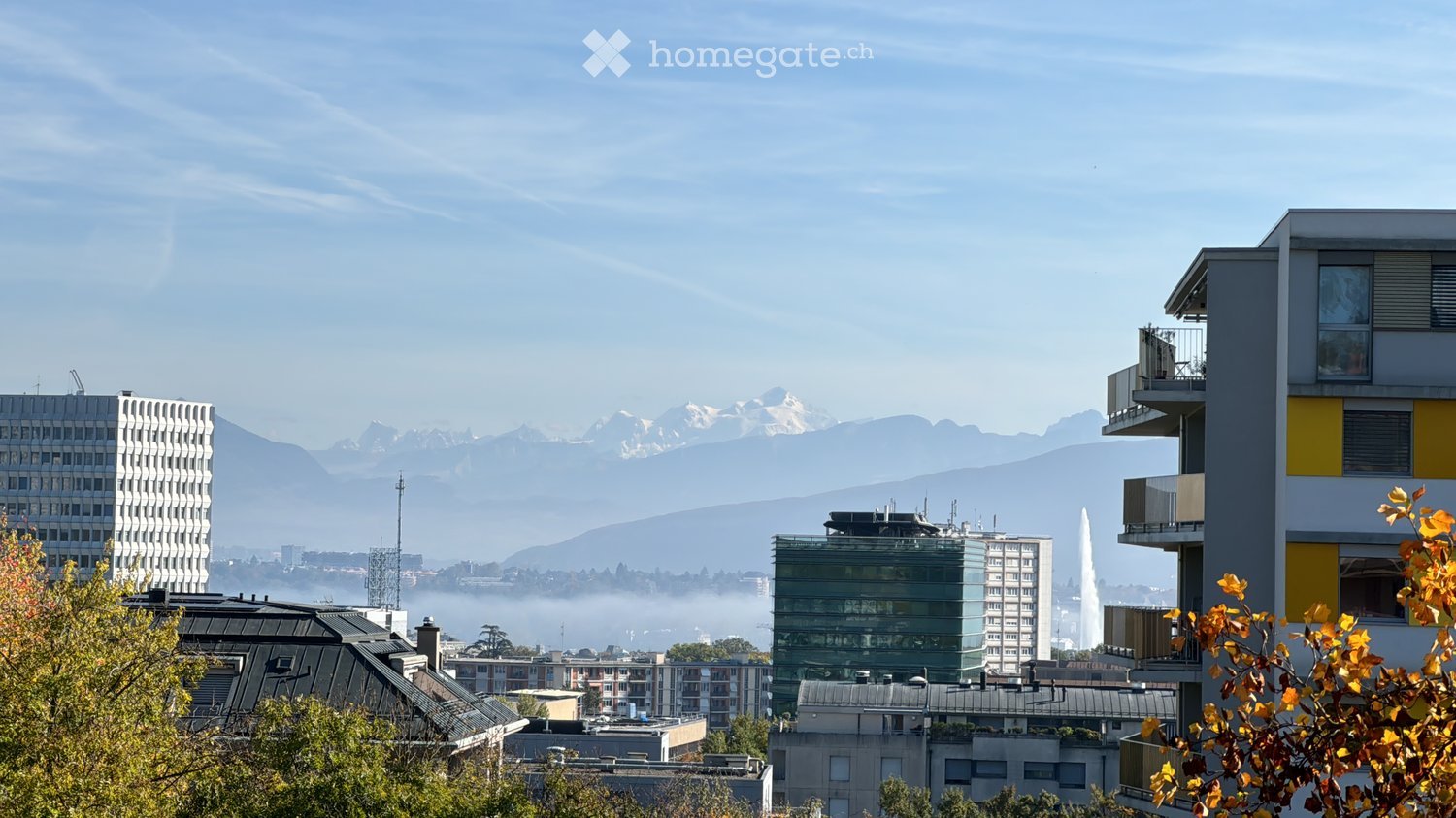 Panoramic view of a cityscape with modern buildings, distant mountains, and a fountain in the foreground.