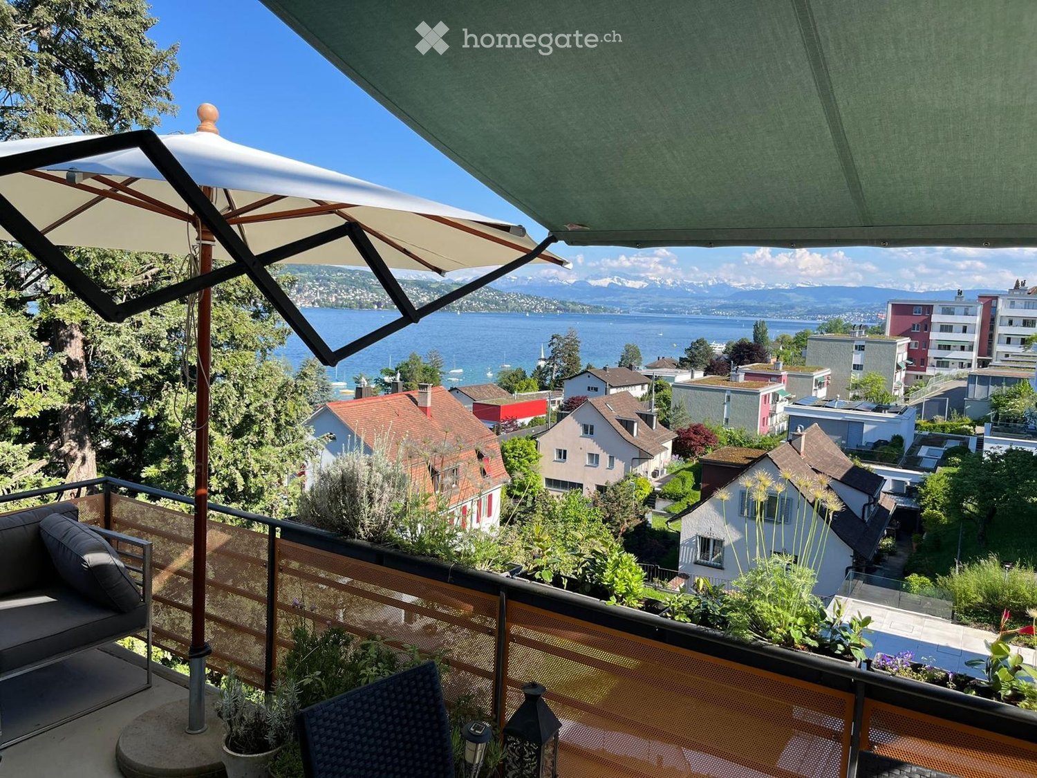Balcony with a view of a lake and surrounding houses, featuring a sofa, chair, table, and umbrella.