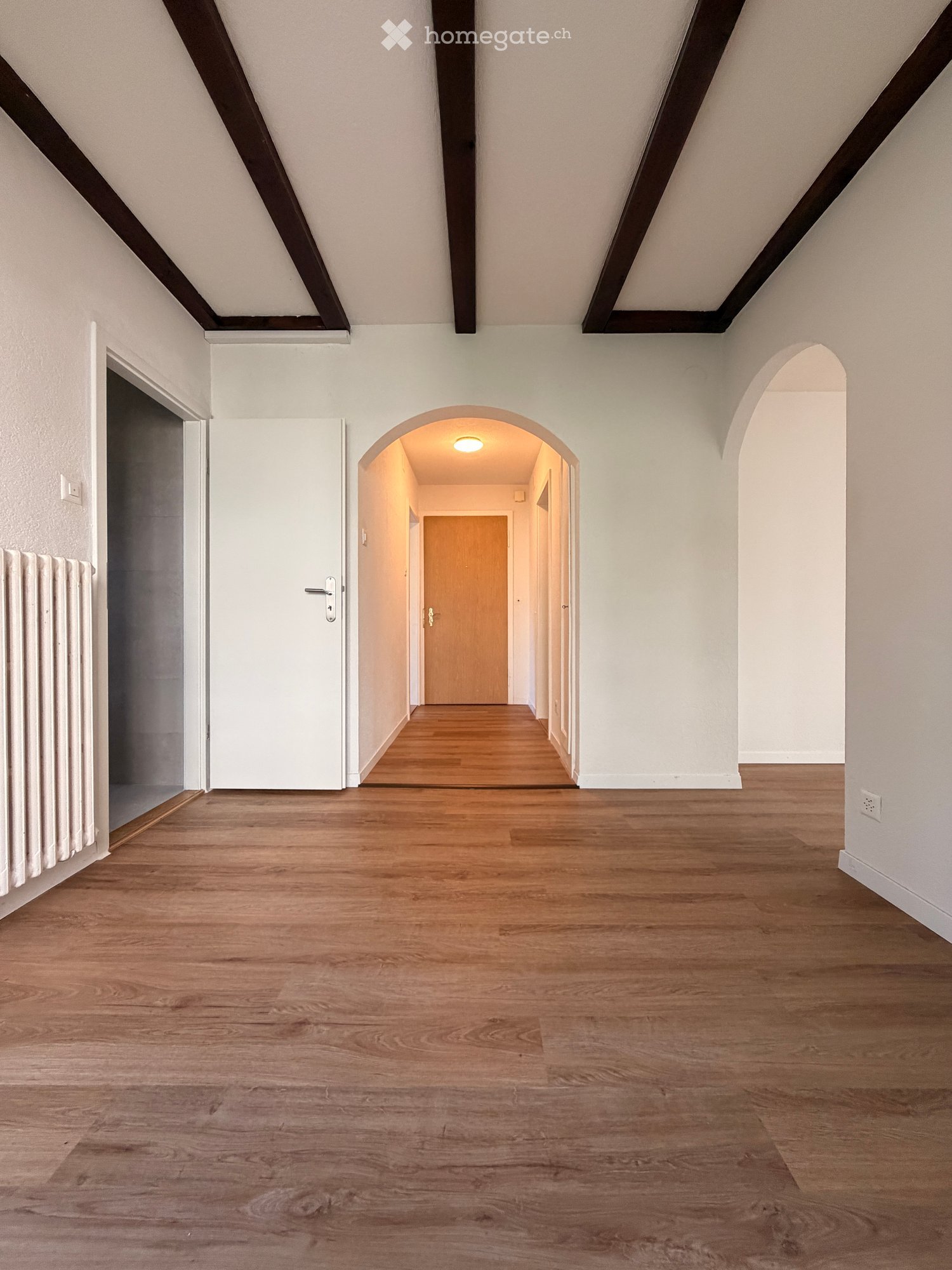Empty hallway with wooden flooring, white walls, and exposed wooden beams on the ceiling. Features an arched doorway leading to another room.