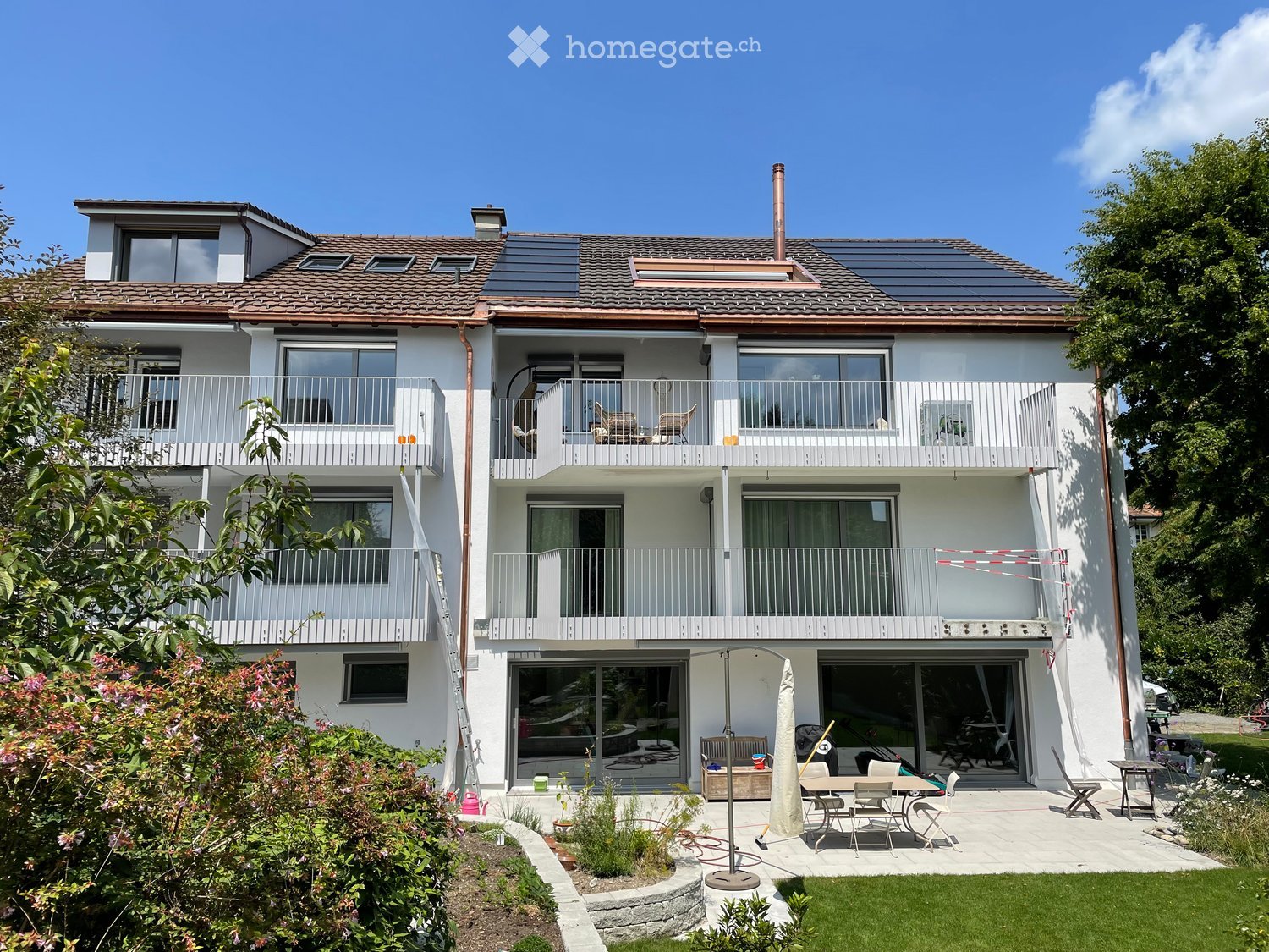 Two-story house with white exterior, multiple balconies, solar panels on the roof, and a well-maintained garden with patio furniture.