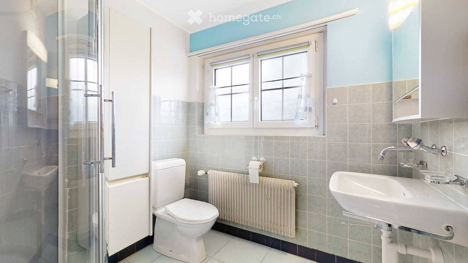 Modern bathroom with white tiles, a toilet, sink, shower, and a window with a curtain. Features a radiator and a mirror above the sink.