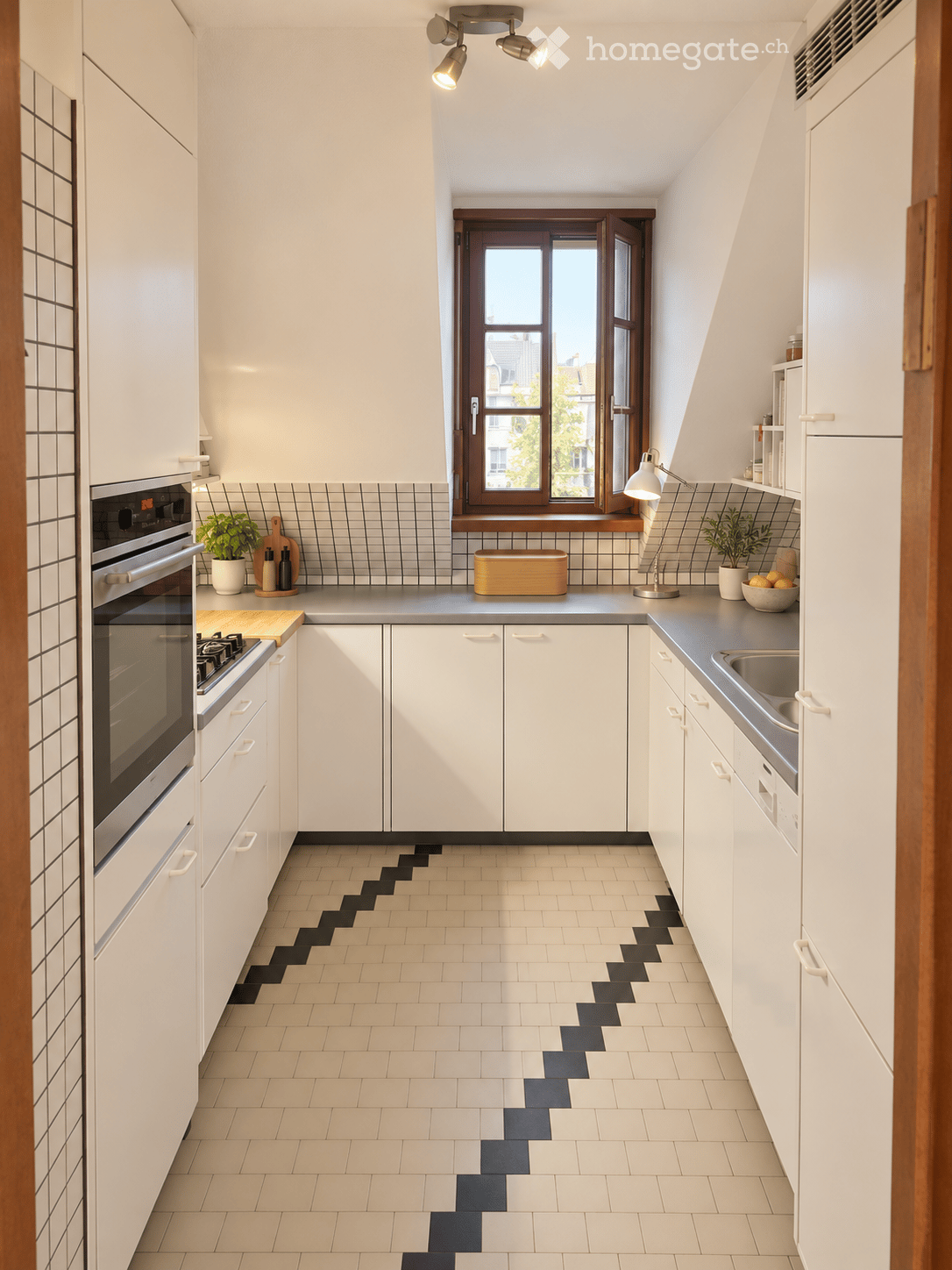 Modern kitchen with white cabinets, stainless steel appliances, and a distinctive black and white tiled floor. Features an oven, sink, countertop, and decorative items including potted plants and a bowl.