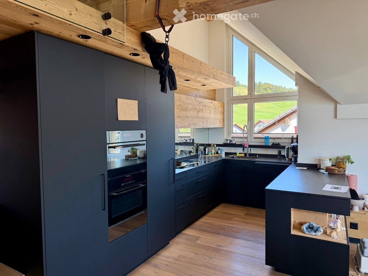 Modern kitchen with dark grey cabinets, wooden flooring, and large windows overlooking a green landscape. Features include an oven, sink, and countertop.