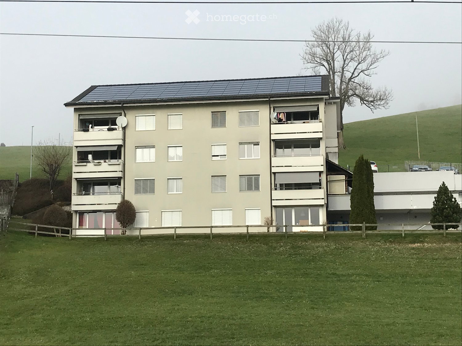 Four-story residential building with balconies, solar panels on the roof, surrounded by a grassy lawn and trees.