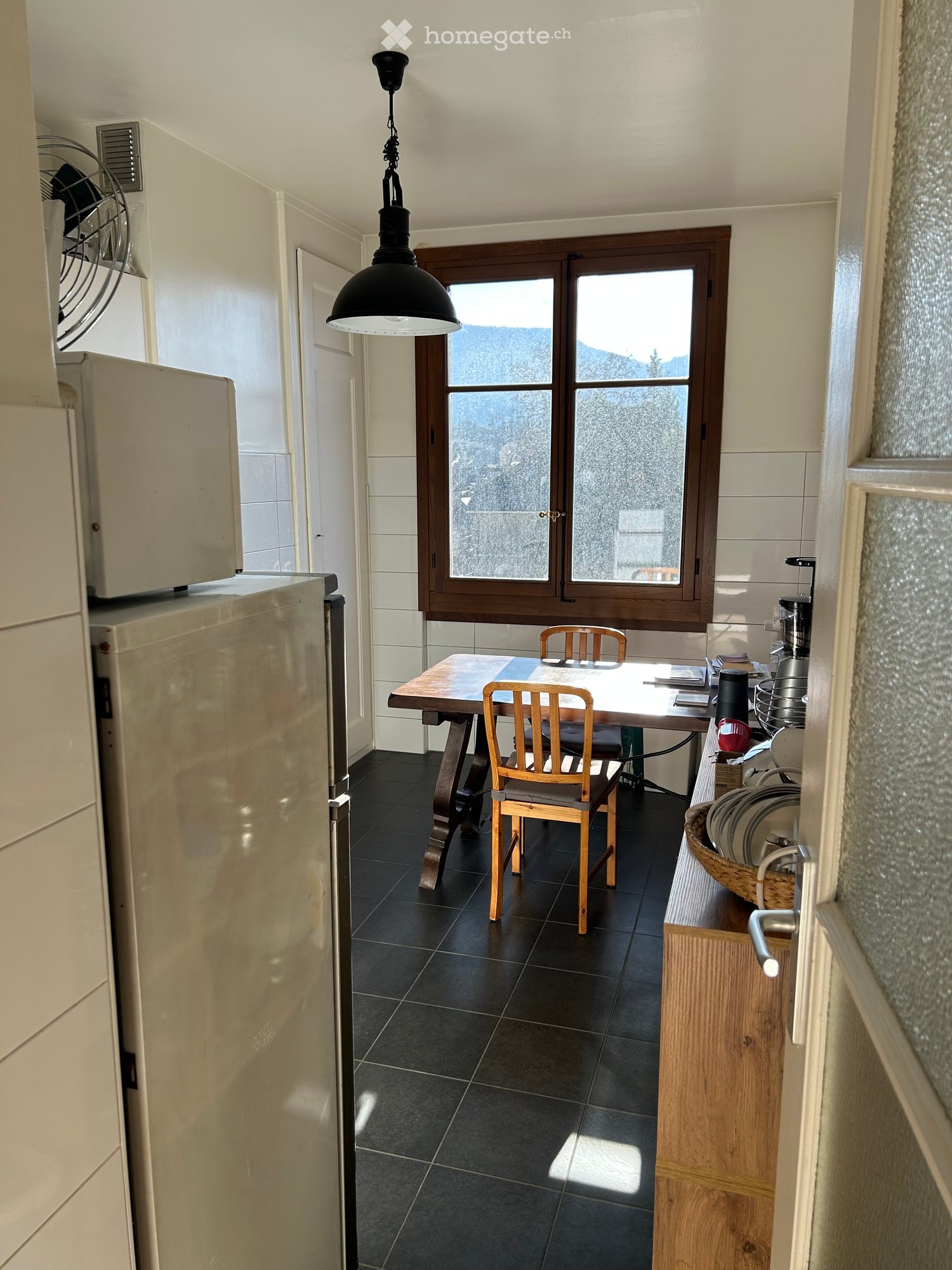 A modern kitchen with dark tiled flooring, a wooden dining table with chairs, a refrigerator, and a window offering a scenic mountain view.