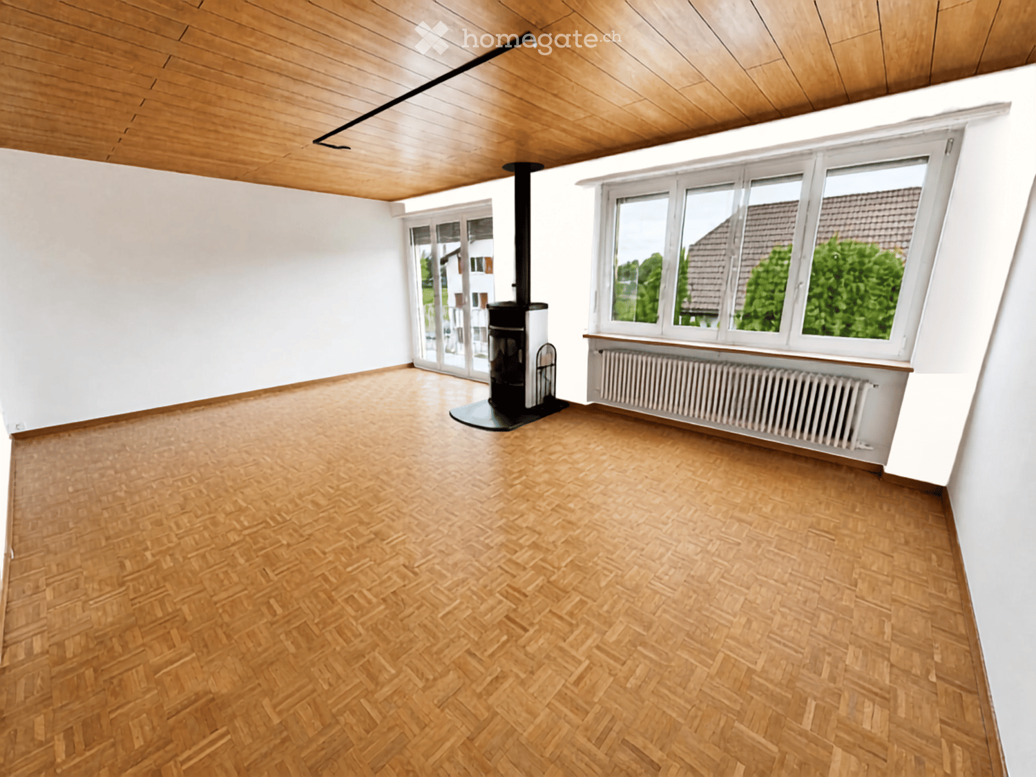 Empty room with wooden herringbone flooring, white walls, and a wood-burning stove. Large windows and a sliding glass door lead to an outdoor area.