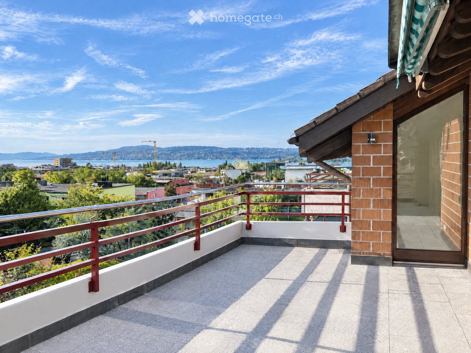 Balcony with a view of a cityscape, lake, and mountains. Features a metal railing, tiled floor, and a glass door leading inside.