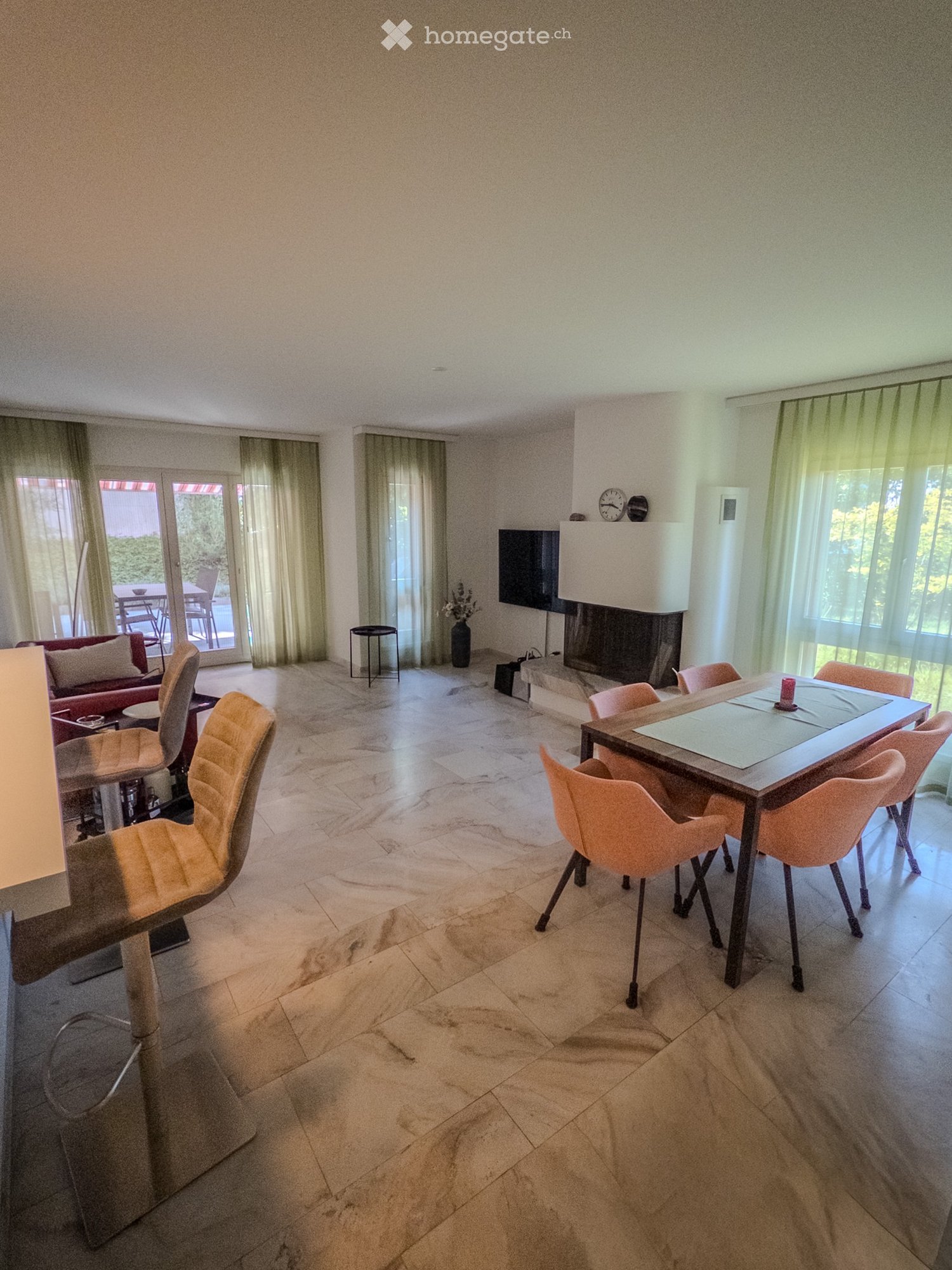Bright living room with marble flooring, a fireplace, a sofa, a dining table with chairs, and a television mounted on the wall. Large windows with sheer curtains let in natural light.