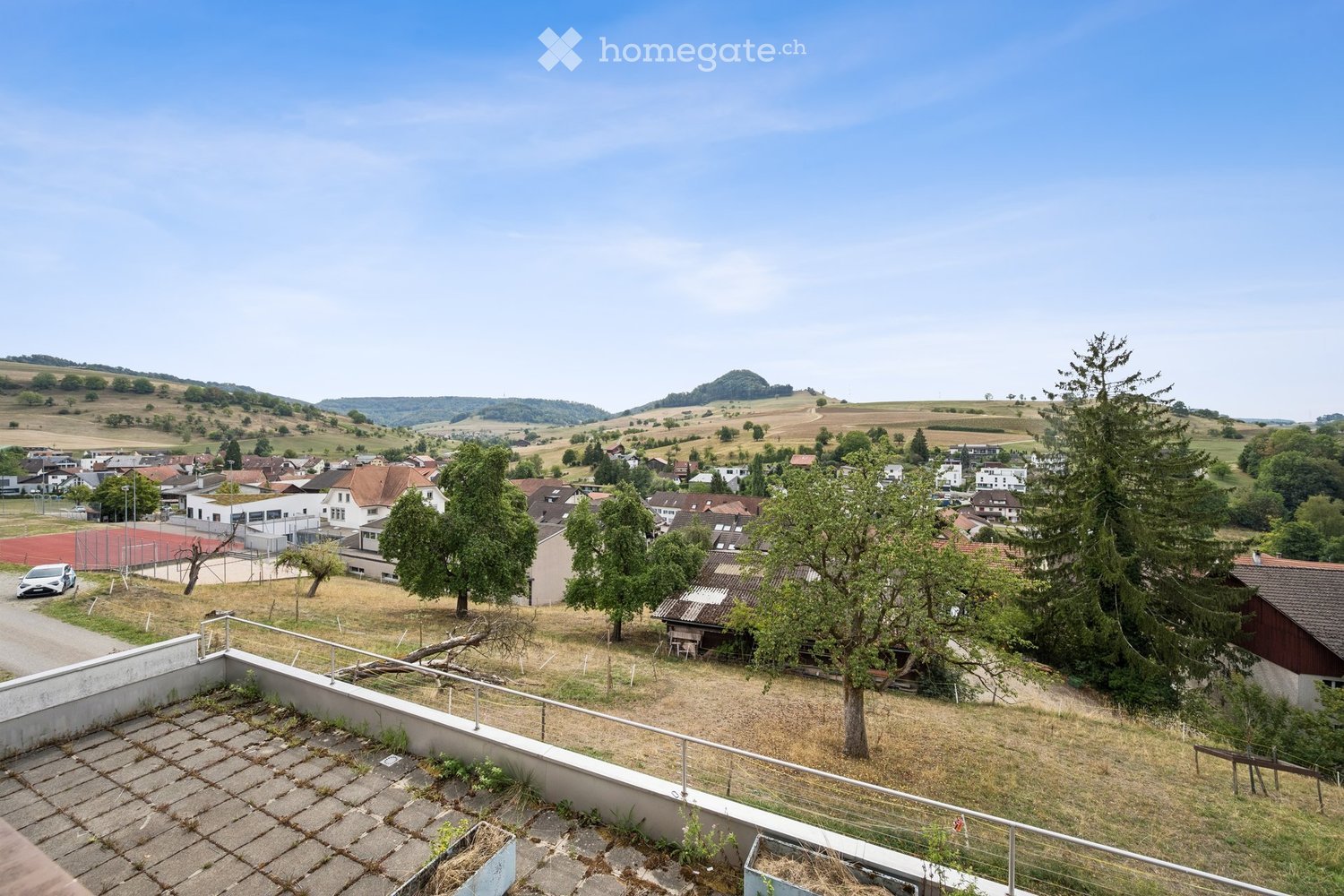 View from a rooftop terrace overlooking a residential area with houses, trees, and a distant landscape.