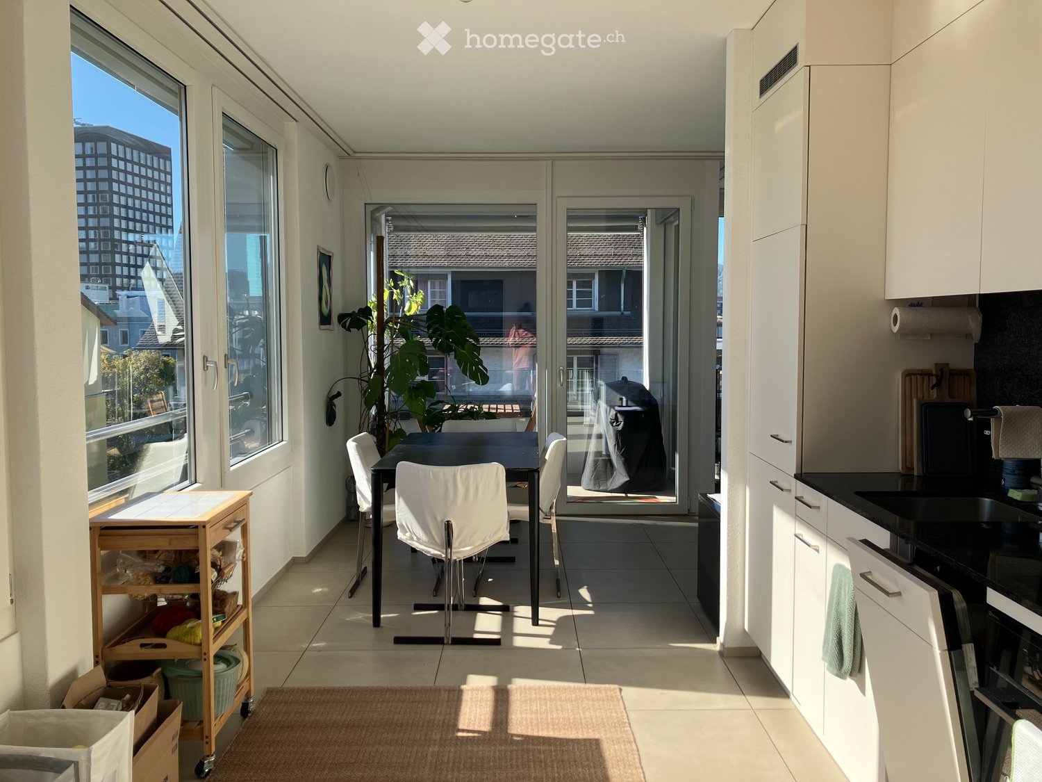 Bright, modern kitchen and dining area with white cabinets, black countertops, a sink, oven, dishwasher, a small cart, a dining table with chairs, and a large plant near the sliding glass doors.