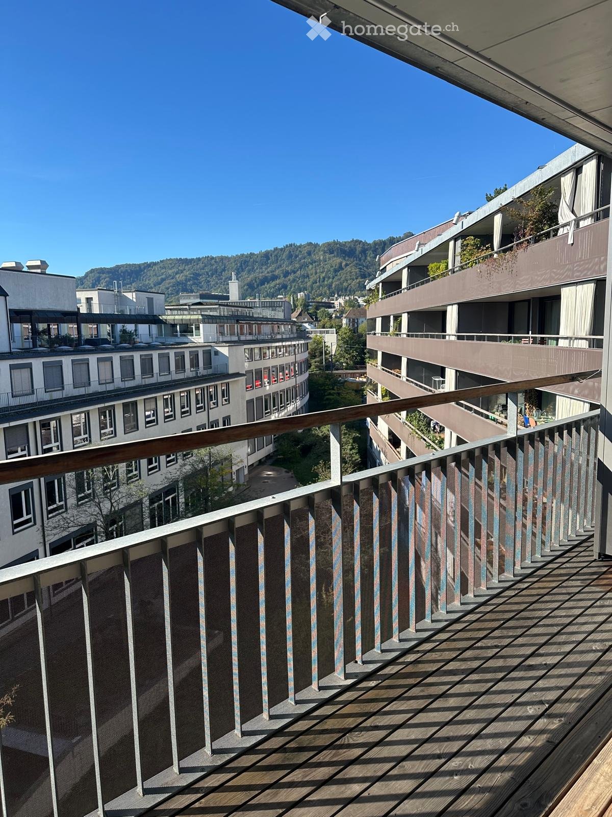 Balcony with wooden decking, metal railing, and view of surrounding buildings and greenery.