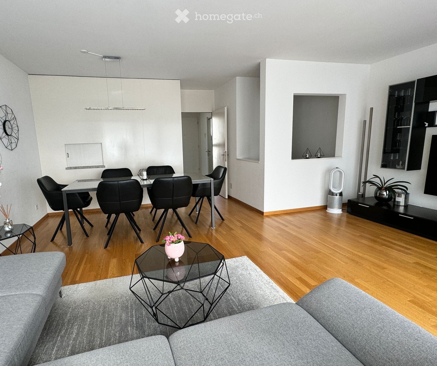 Modern living room with grey sofa, glass coffee table, dining area with black chairs and table, TV stand, and decorative items.