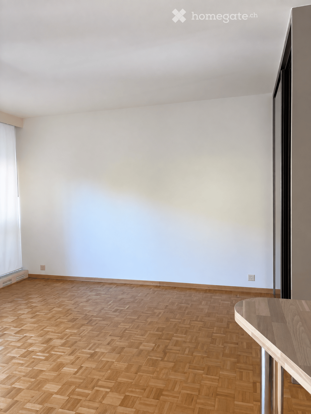Empty room with light grey walls and herringbone wooden flooring, featuring a large window with blinds and a small wooden table in the corner.