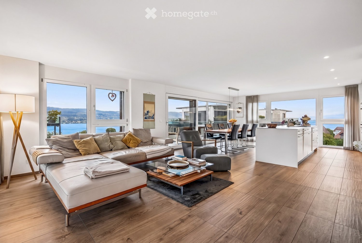 Bright, open-plan living and dining area with large windows offering a scenic view, featuring a white leather sofa, coffee table, dining table, and modern kitchen island.
