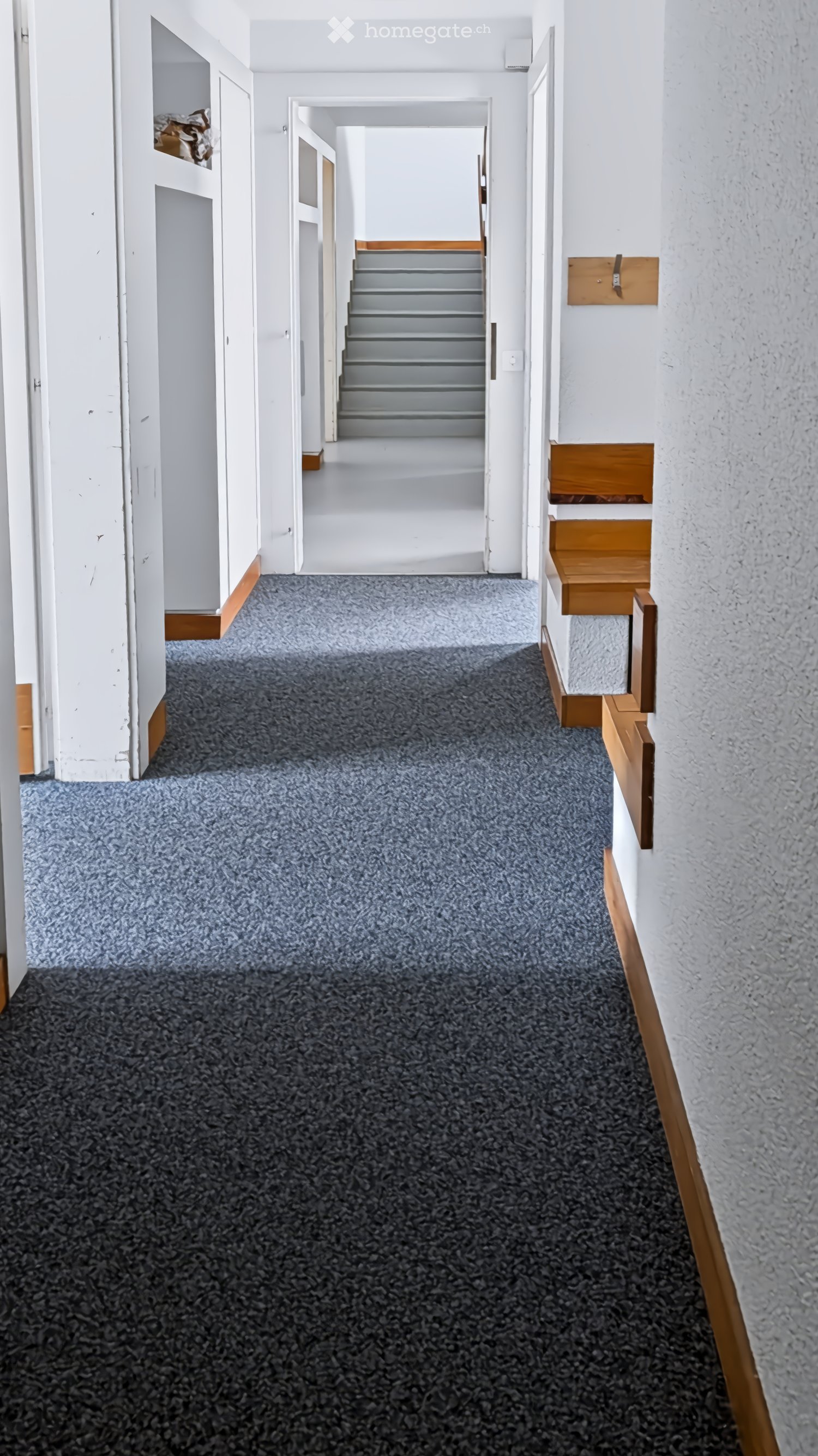 A clean, carpeted hallway with white walls, wooden trim, and a staircase leading upwards.