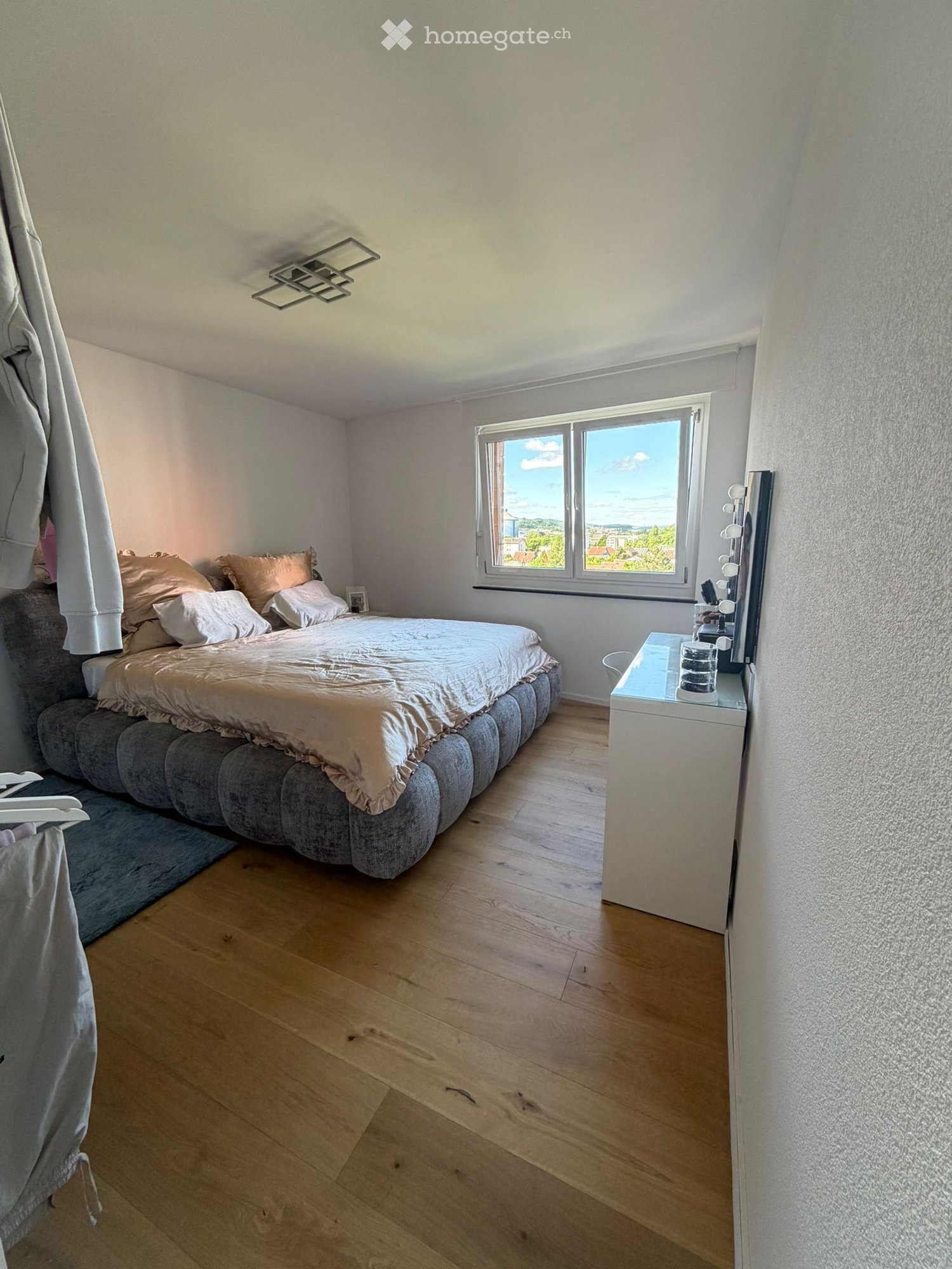 Bedroom. Cozy bedroom with wooden flooring, large window offering a view, a grey upholstered bed with pillows and a duvet, a nightstand with a lamp and mirror, and a small rug near the bed.