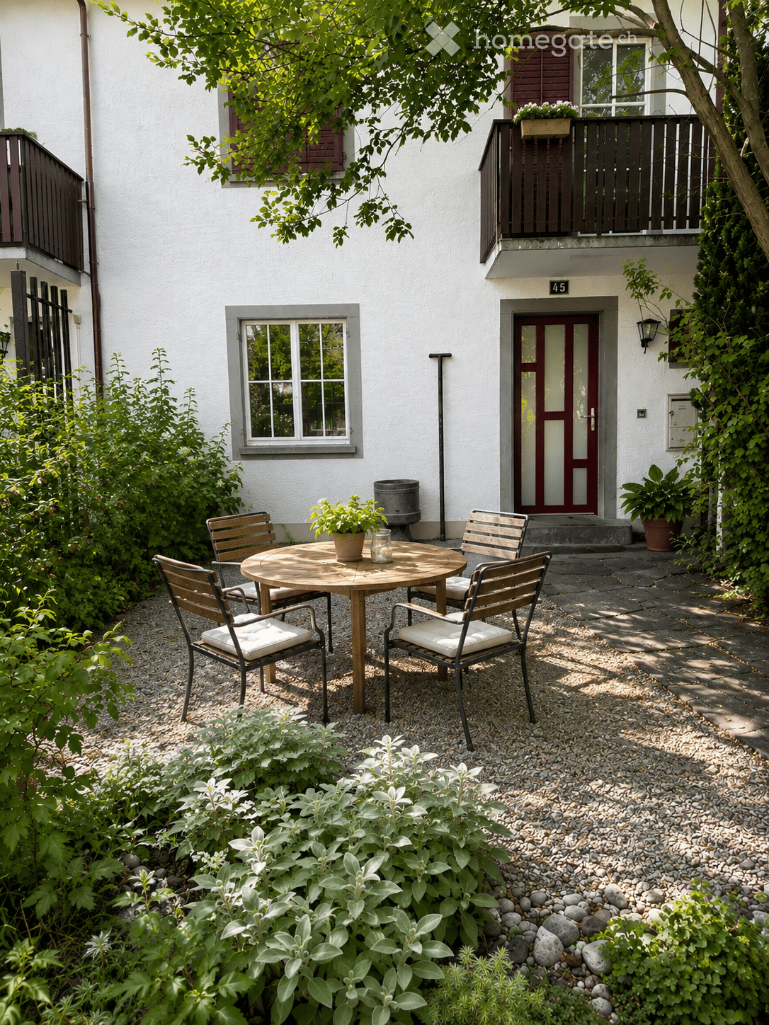 A small, well-maintained garden area with a round wooden table and four matching chairs, surrounded by greenery and a gravel pathway.
