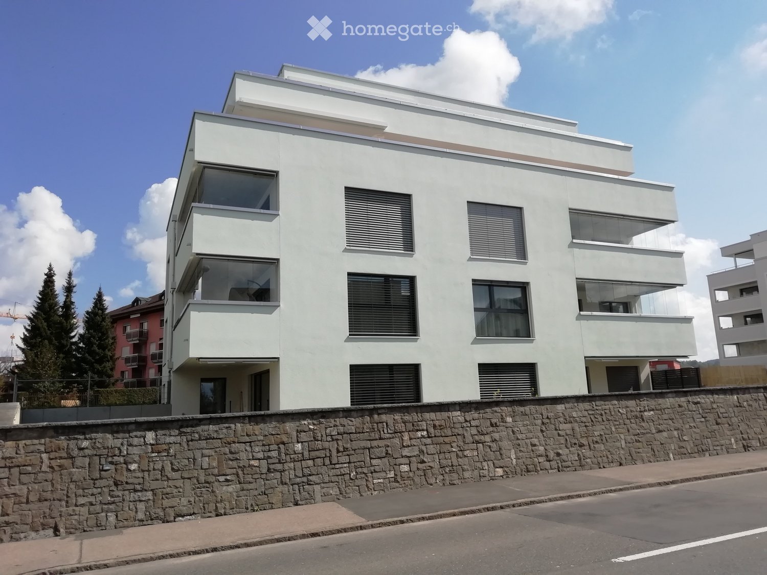 Modern, light-colored building with large windows and balconies, situated on a raised ground floor with a stone wall in front.