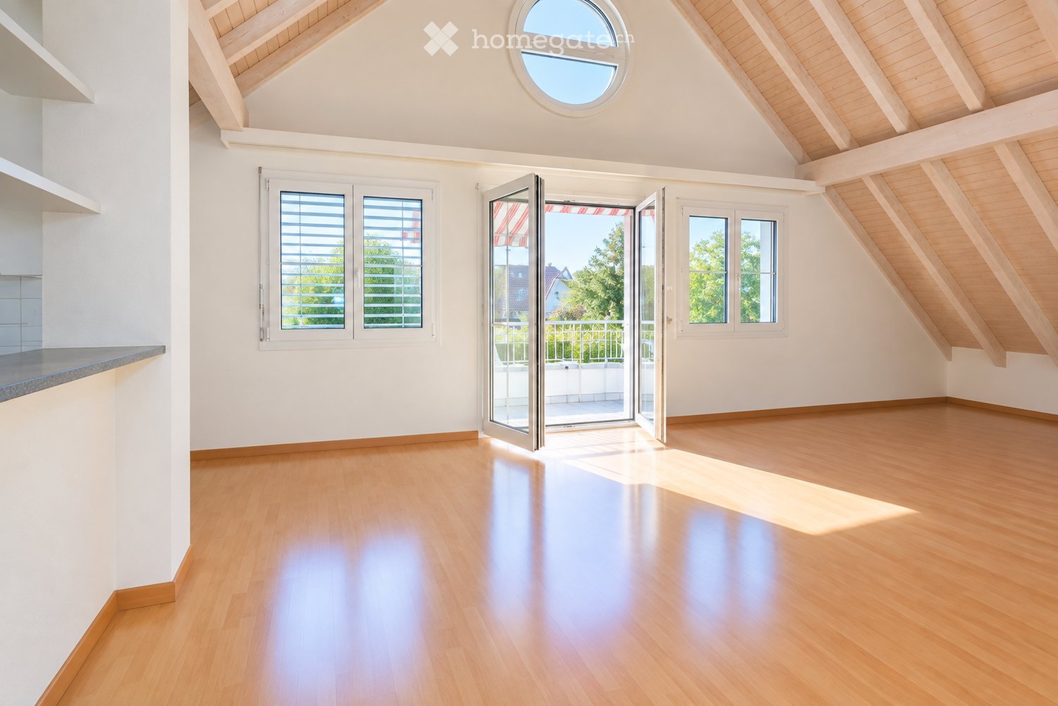 Bright, empty room with wooden flooring, white walls, and large windows, including a skylight and sliding glass doors leading to a balcony.