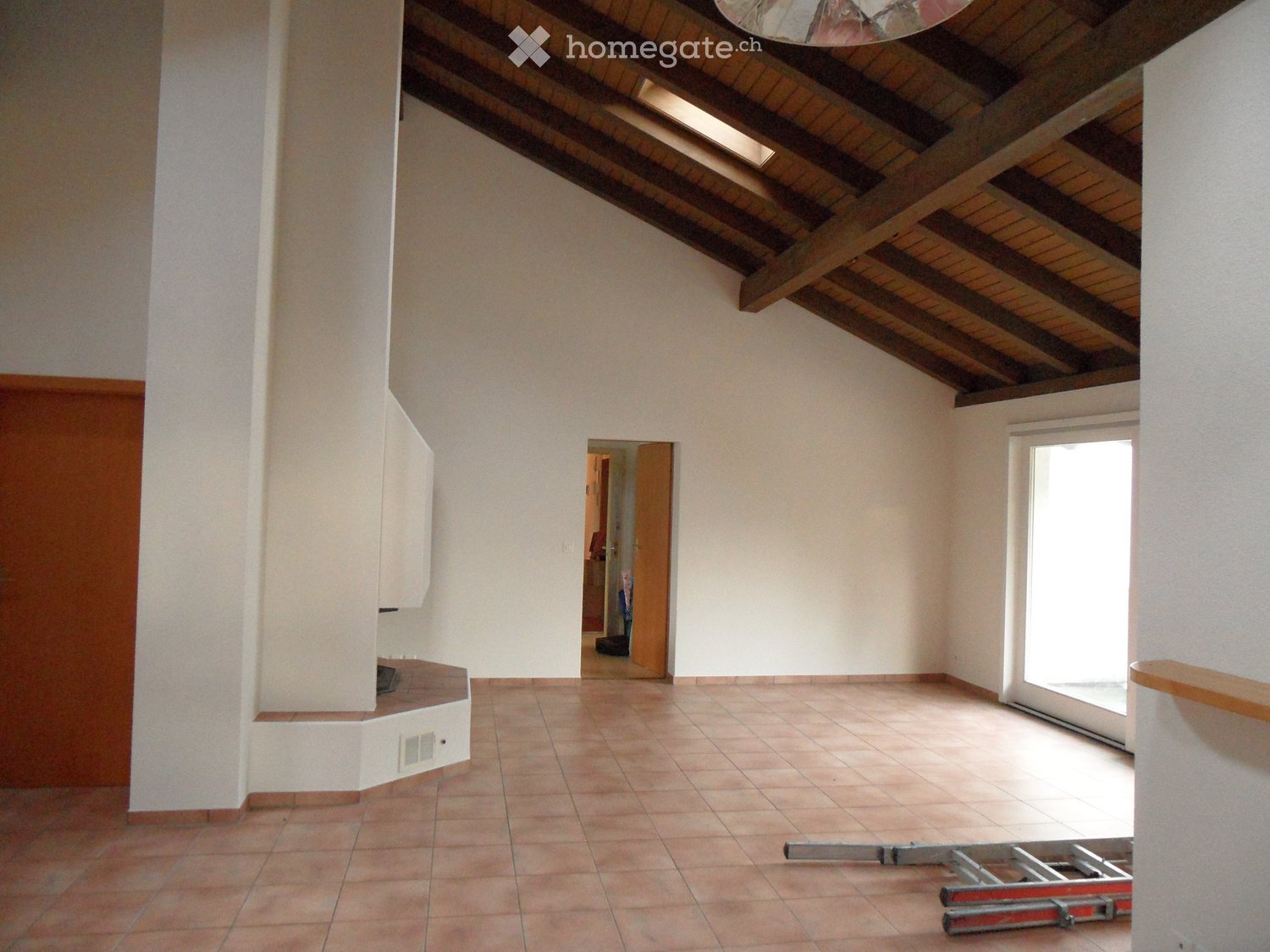 Empty room with exposed wooden beams on the ceiling, tiled floor, and a large window letting in natural light. There is a doorway leading to another room and some construction equipment visible.