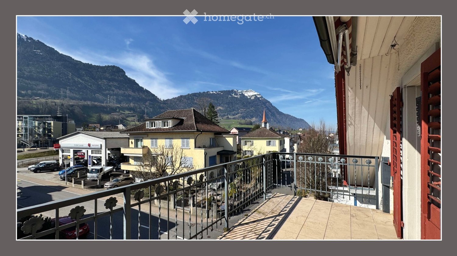 Balcony overlooking a town with mountains in the background, featuring a metal railing and tiled floor.