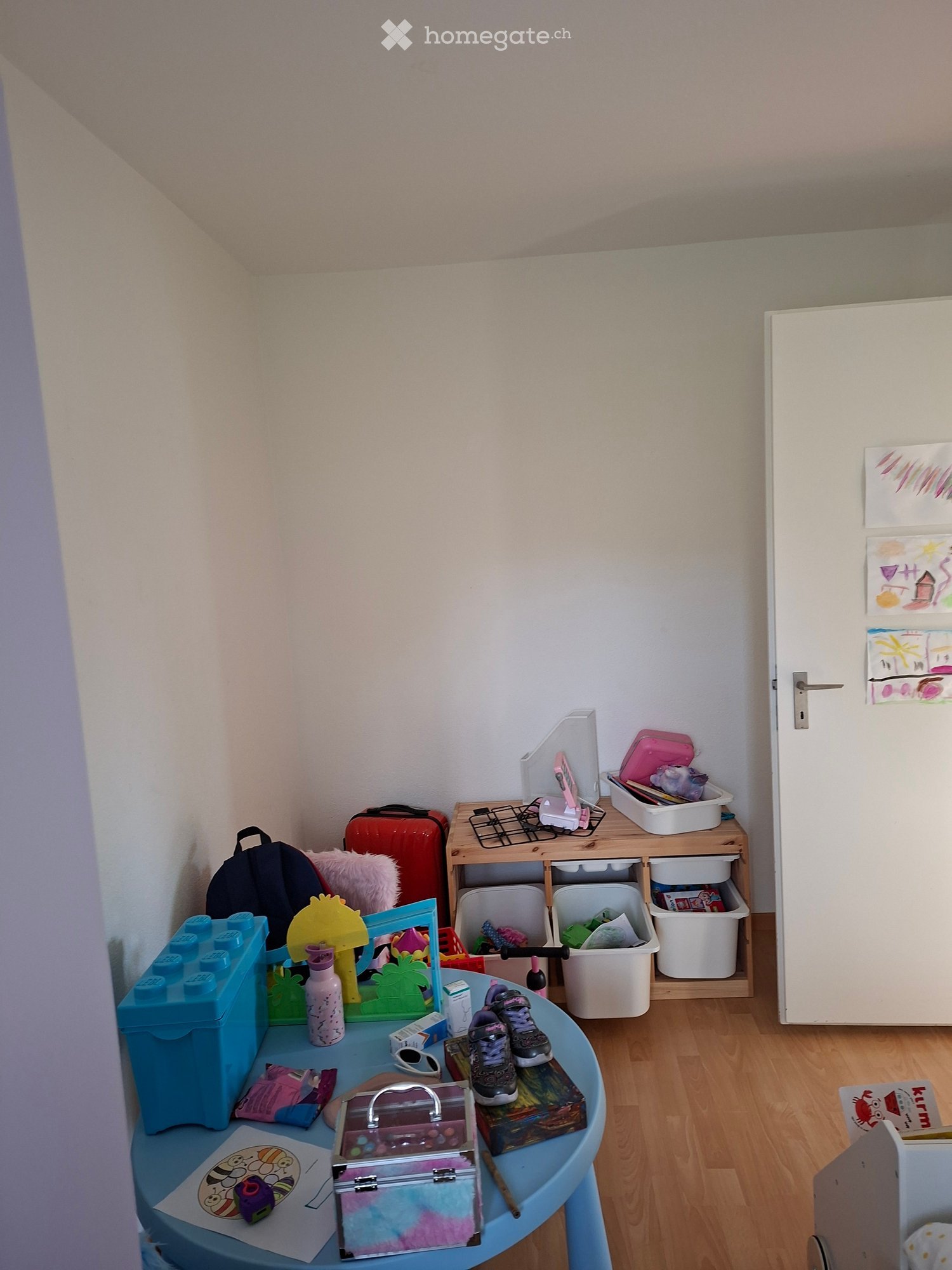 A small, cozy room with light-colored walls and wooden flooring. A blue table holds various items including a toy, a bottle, a cup, and shoes. A storage unit with drawers and shelves is positioned against the wall, containing toys and other items. A backp