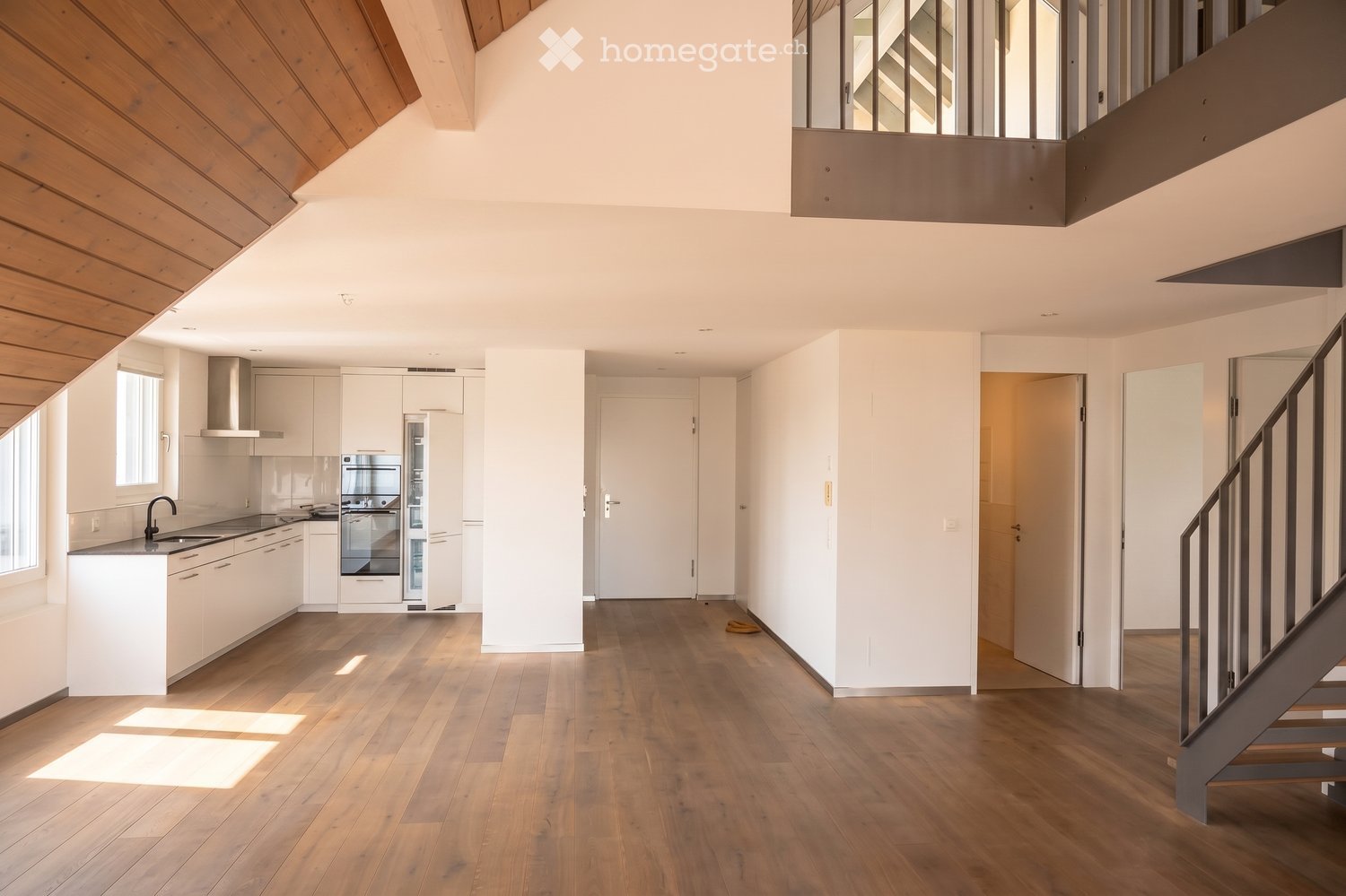 Modern, open-plan living space with a kitchen area featuring white cabinets, stainless steel appliances, and a wooden floor. A staircase leads to an upper level with a wooden slanted ceiling.