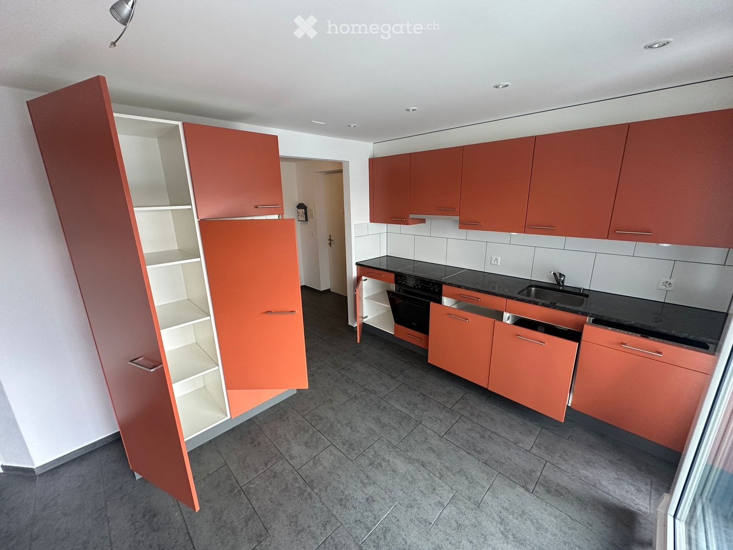Modern kitchen with red cabinets, black countertops, and stainless steel appliances. Open shelving unit on the left side.