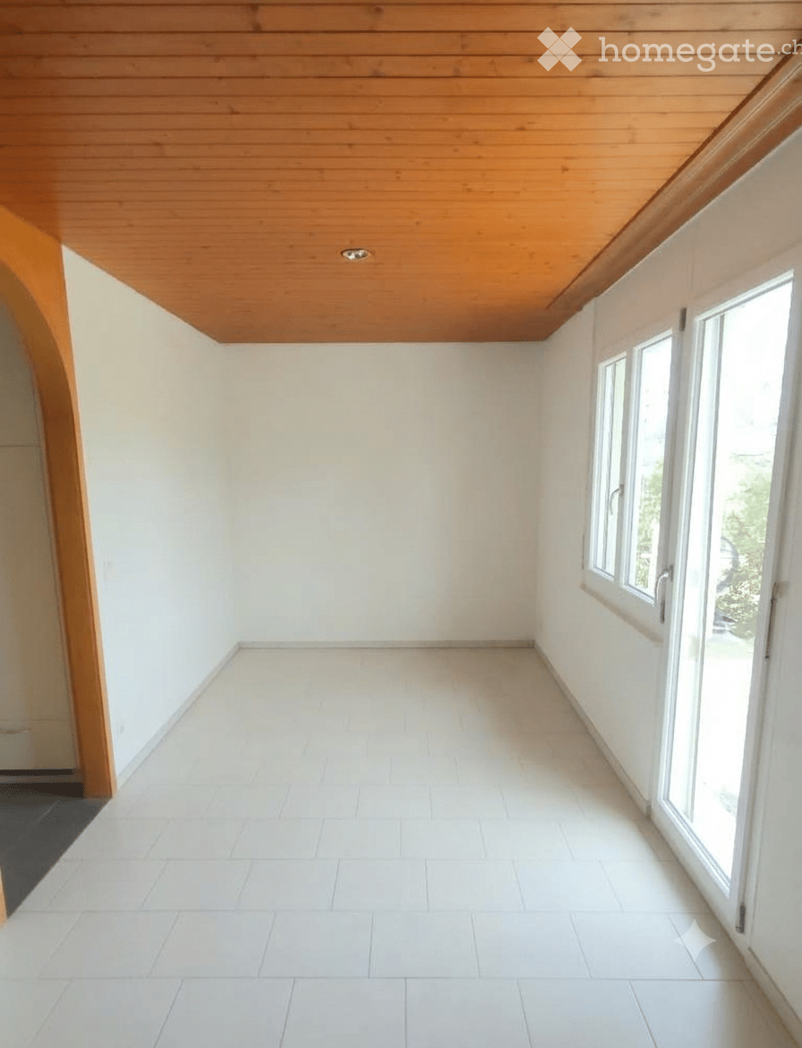 Empty room with white tiled floor, white walls, wooden slanted ceiling, and large windows on one side.