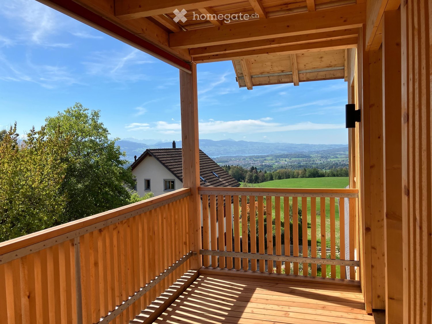 Wooden balcony with railing, overlooking a scenic valley and mountains under a clear blue sky.