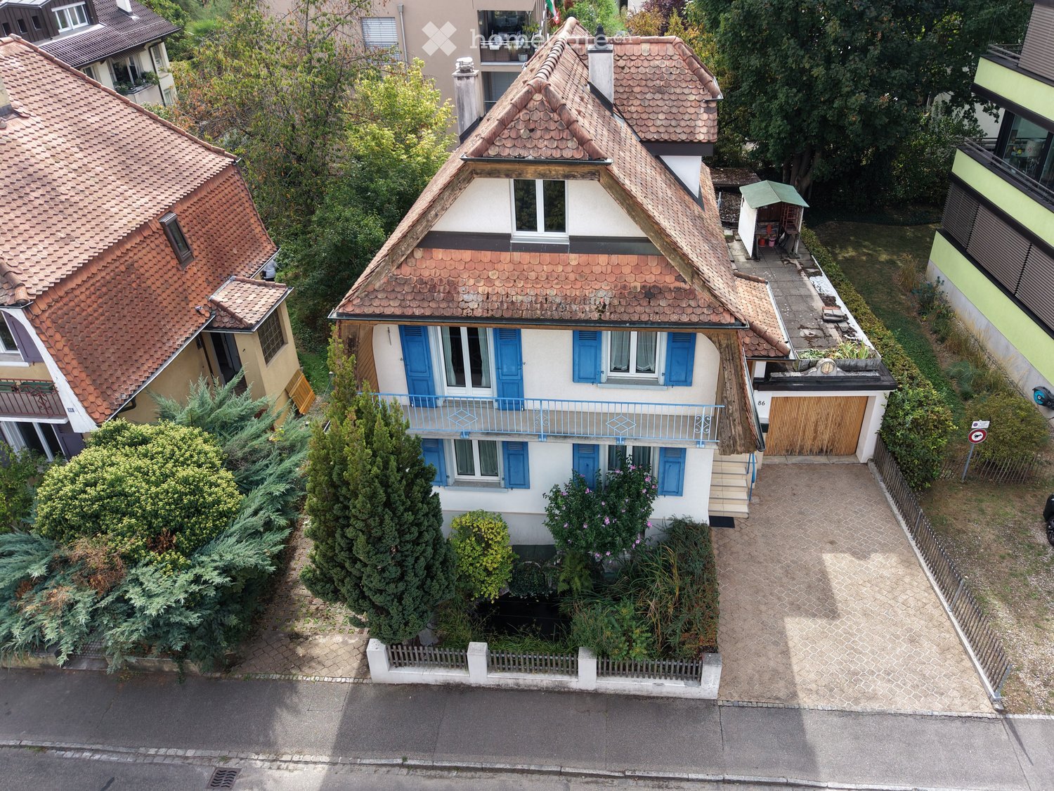 A two-story house with a red-tiled roof, white walls, and blue shutters. The house features a small balcony on the upper floor and is surrounded by greenery and neighboring houses.