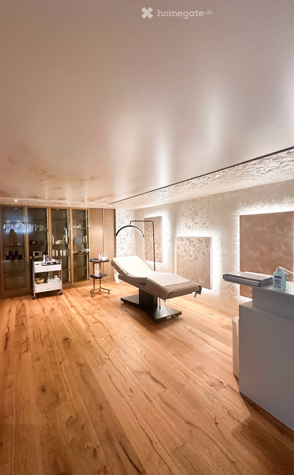A modern, well-lit treatment room with wooden flooring, a treatment bed, a lamp, a small table, and a cabinet with shelves.