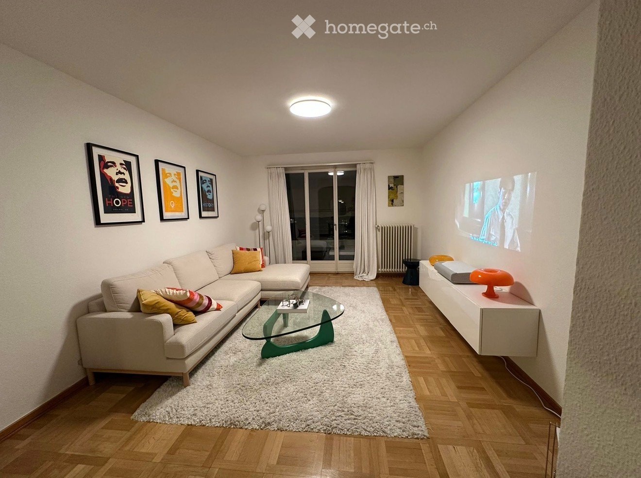 Modern living room with a white sectional sofa, glass coffee table, white shag rug, and large screen projection on the wall. The room features wooden flooring, large windows with curtains, and decorative picture frames.