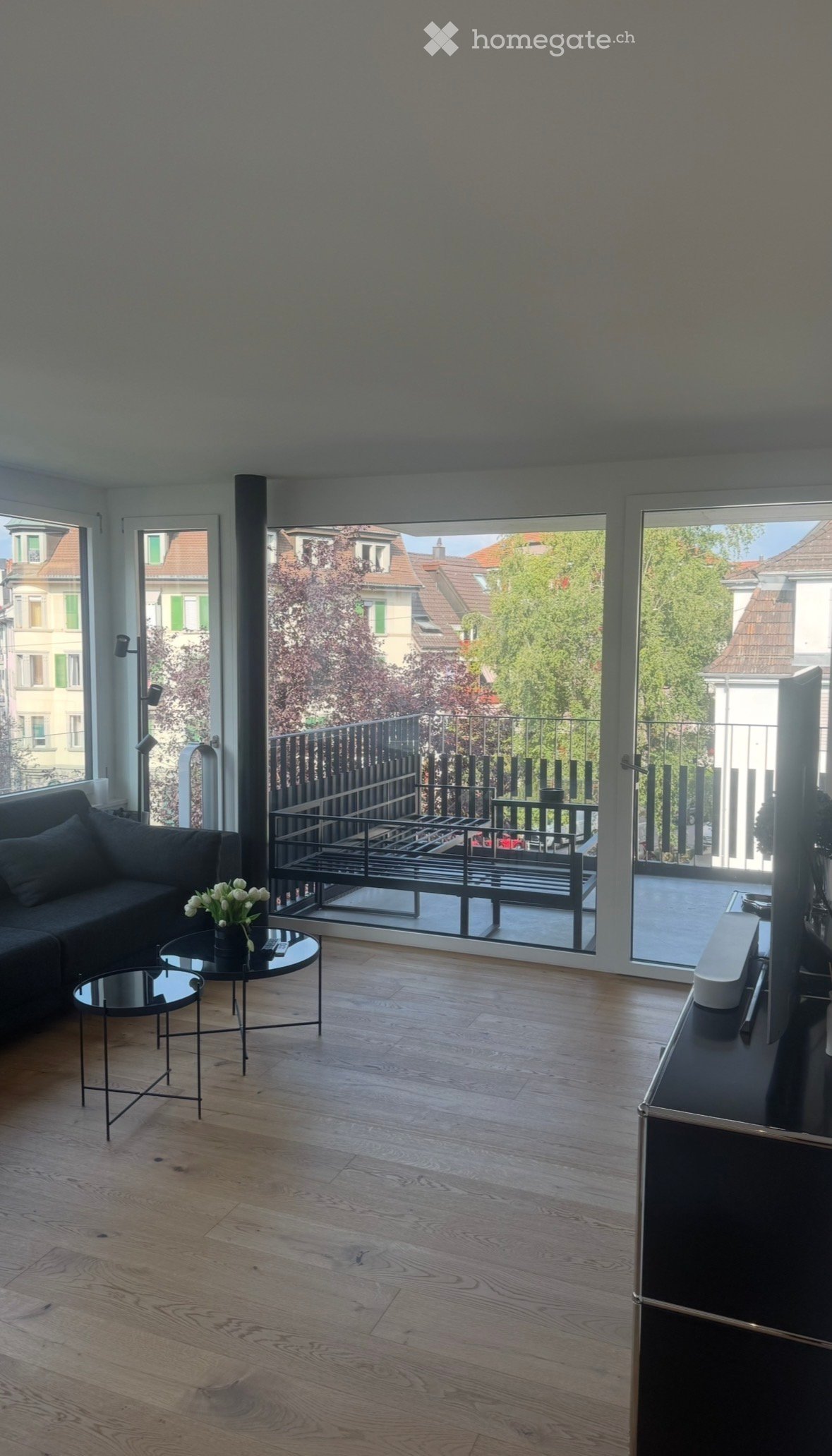 Modern living room with wooden flooring, large windows, balcony access, sofa, coffee table, small round table, vase with flowers, and TV screen.