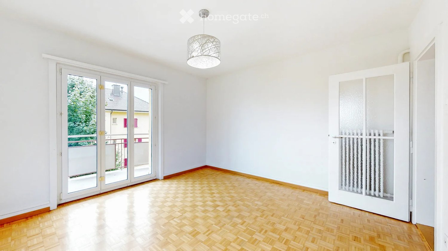Empty room with wooden flooring, white walls, large glass sliding doors leading to a balcony, and a modern hanging light fixture.