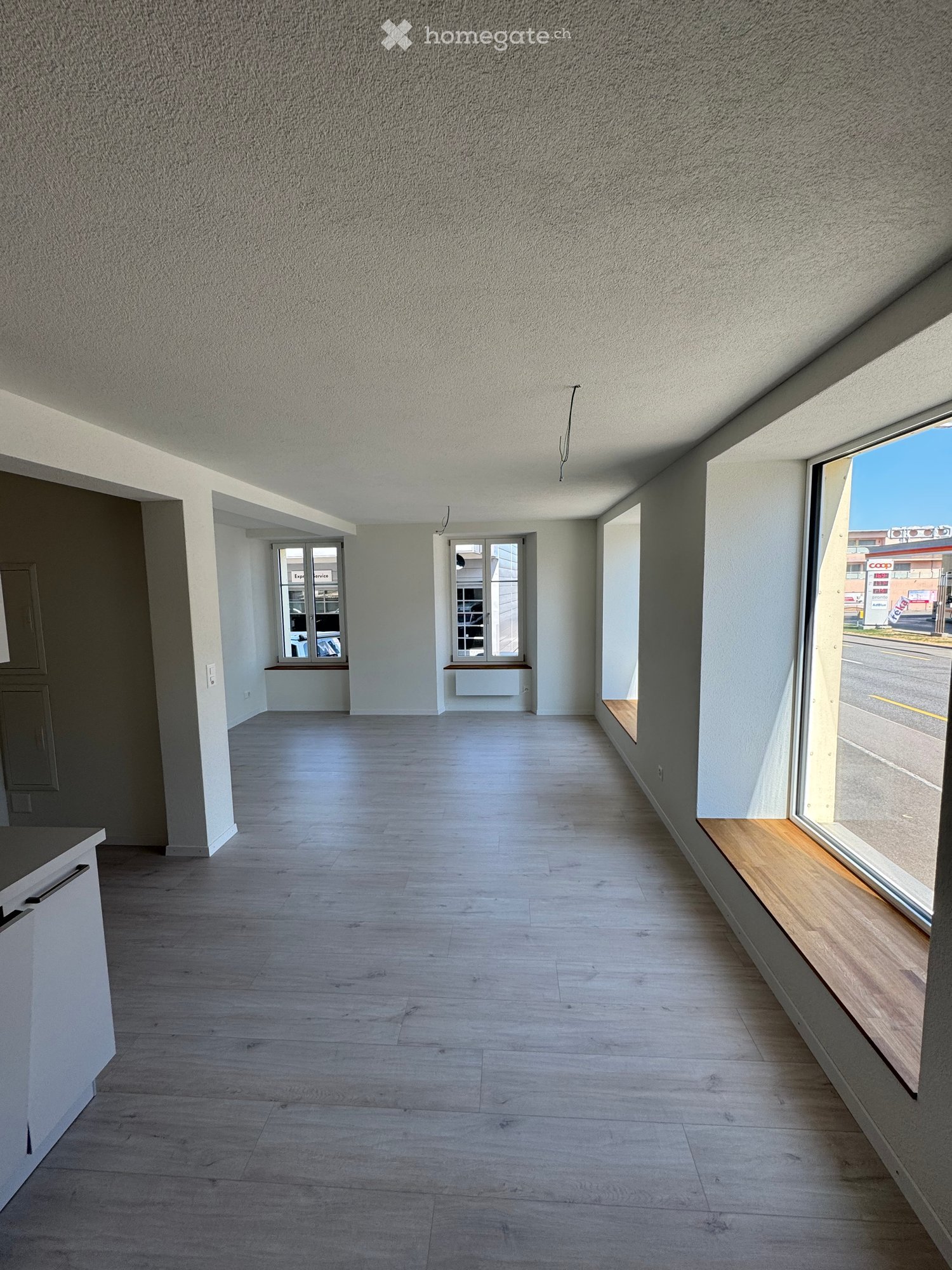 Empty room with light wooden flooring, white walls, and large windows allowing natural light. There are small built-in benches near the windows and a small cabinet on the left side.