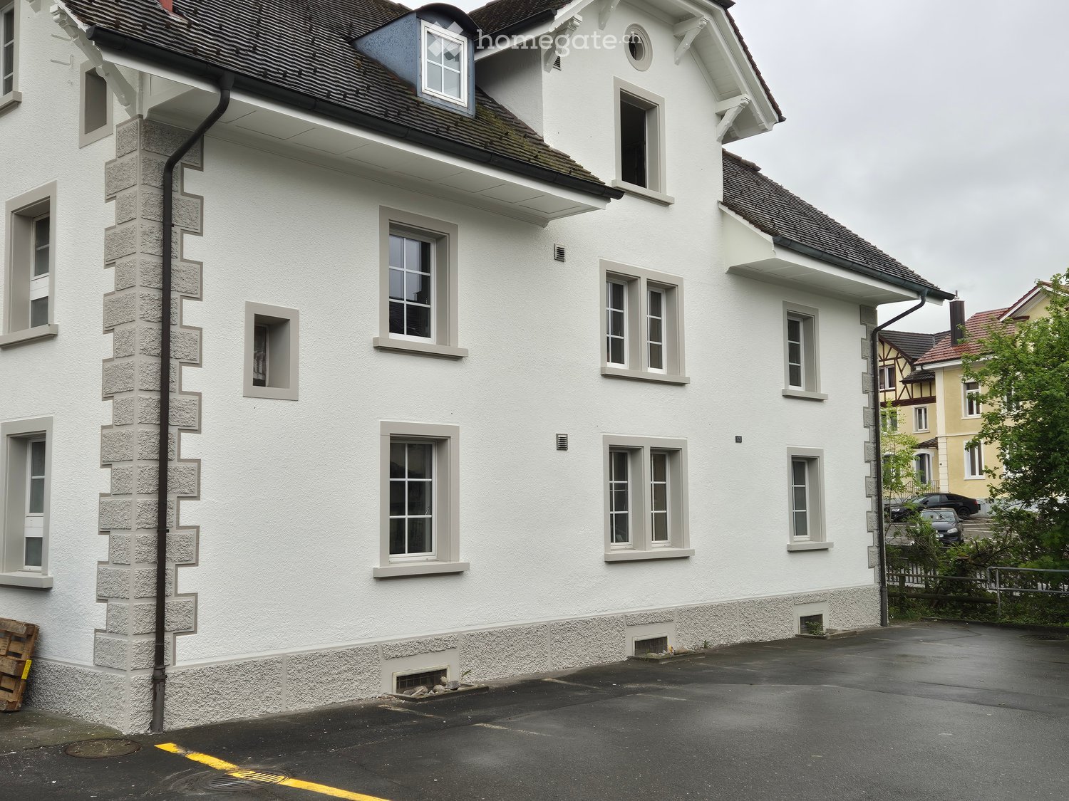 Two-story white building with multiple windows and a dark roof, featuring a stone base and a small balcony on the upper floor.