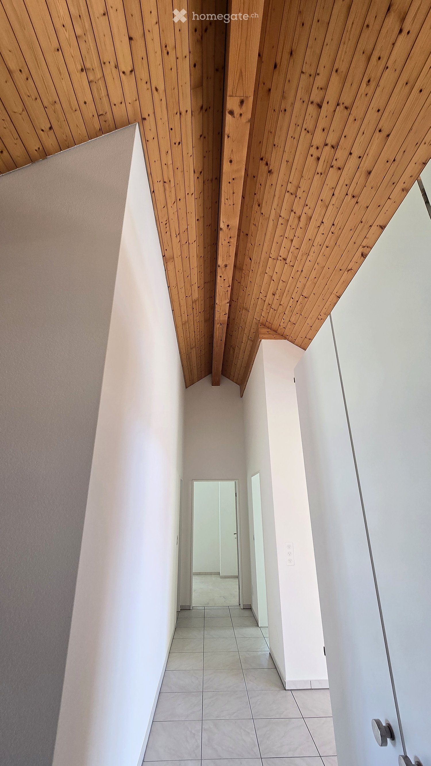 Modern hallway with white walls, tiled floor, and wooden slatted ceiling. Features a mirror and a light switch on the wall.