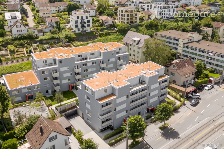 Aerial view of a modern multi-story residential building complex with grey facades, orange-tiled roofs, and surrounding greenery.