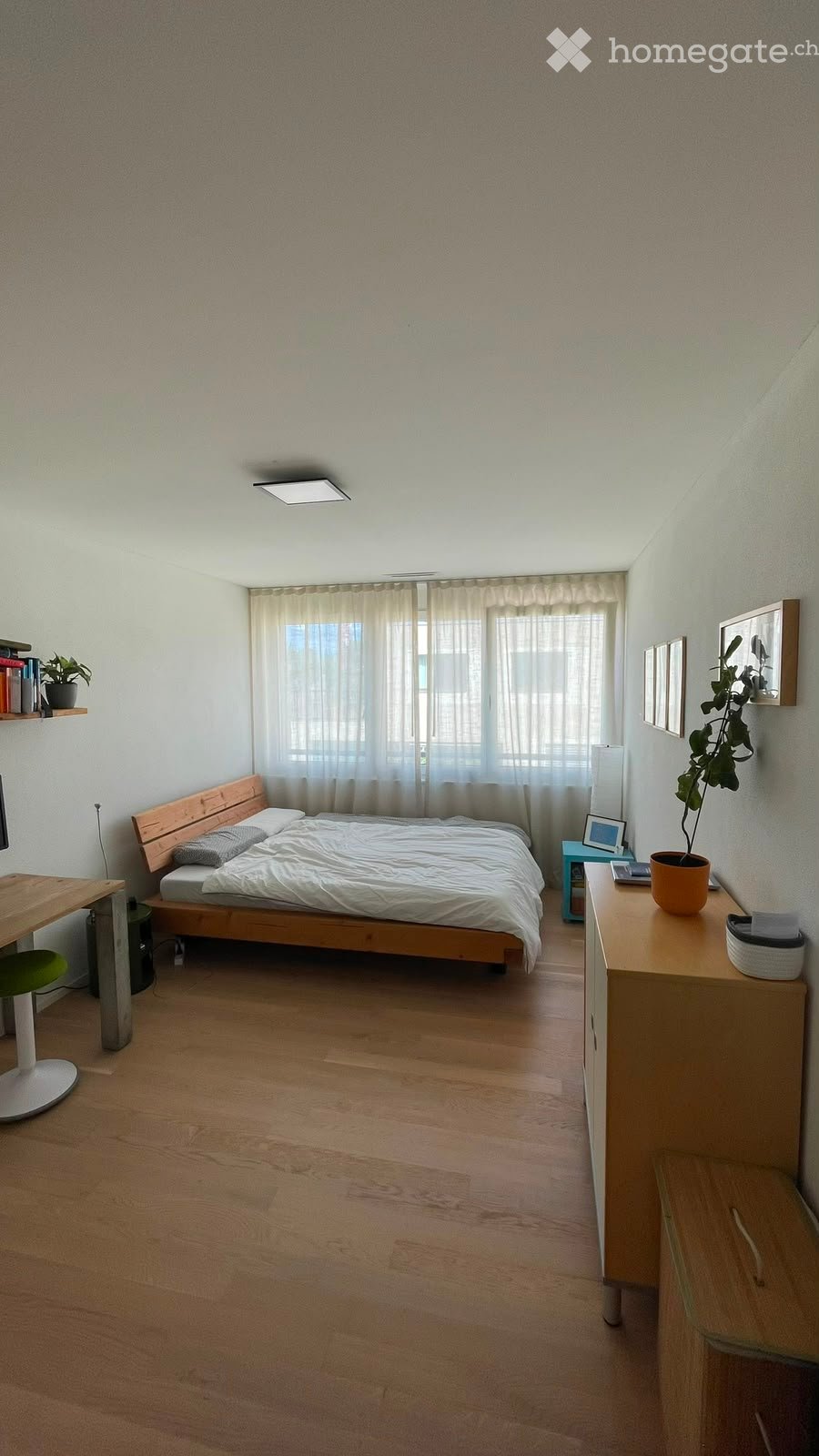Cozy bedroom with wooden flooring, a bed with white bedding, a nightstand with a lamp, a desk with a chair, and a shelf with books and a plant.