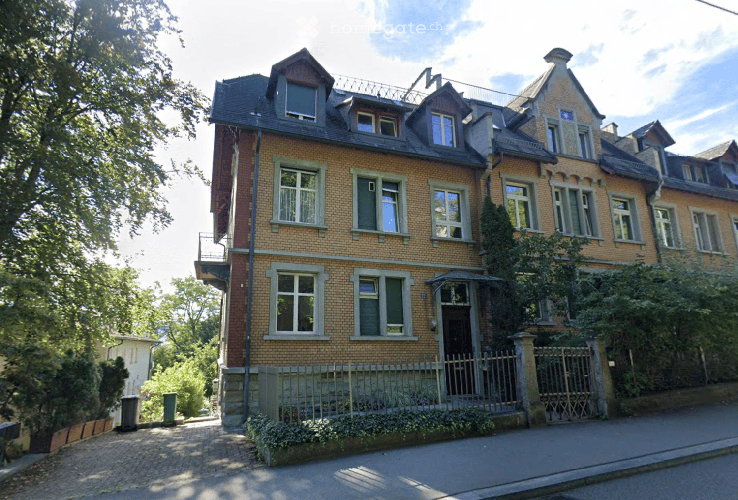 A multi-story brick house with multiple windows, a balcony, and a gated entrance, surrounded by greenery.