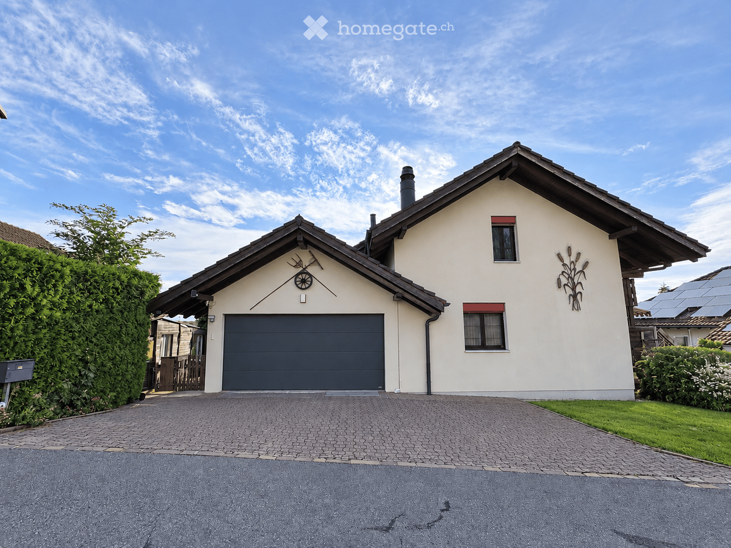 Two-story house with a gray garage door, white walls, and decorative corn stalk wall art. The house features a paved driveway and is surrounded by greenery.