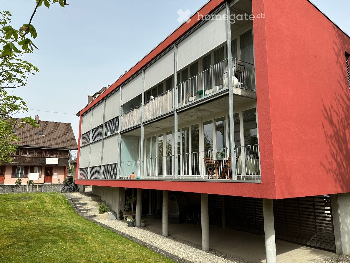 2-story apartment building with a red exterior, large windows, and balconies on the upper floor. The building is surrounded by a grassy area and other residential structures.