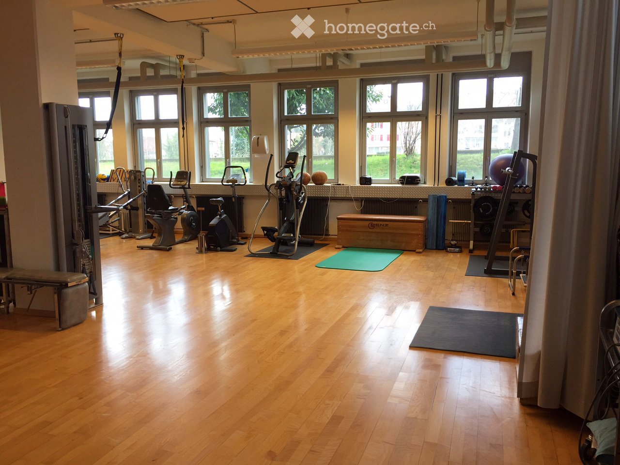 Well-lit fitness room with wooden flooring, large windows, and various exercise equipment including bikes, a treadmill, weights, and yoga mats.