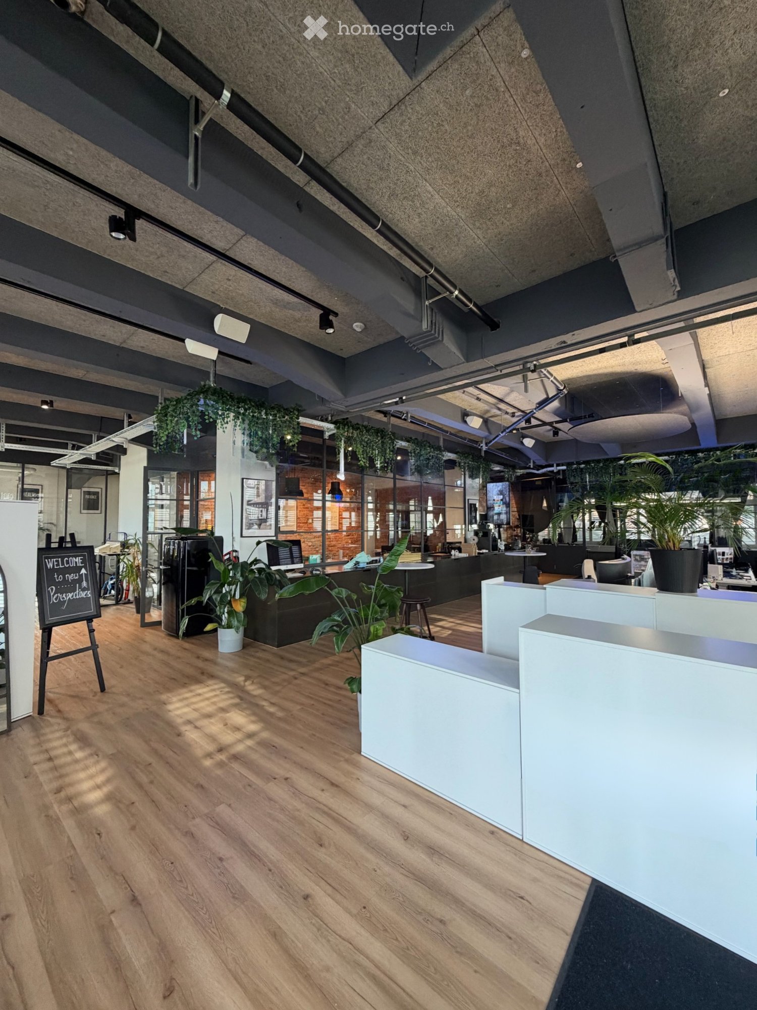 Modern office space with wooden flooring, white partitions, desks, chairs, and plants. A welcome sign is visible on the left side.