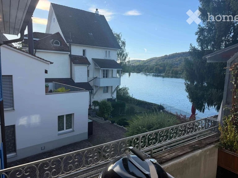 Balcony overlooking a serene lake with surrounding greenery and a multi-story house in the background.