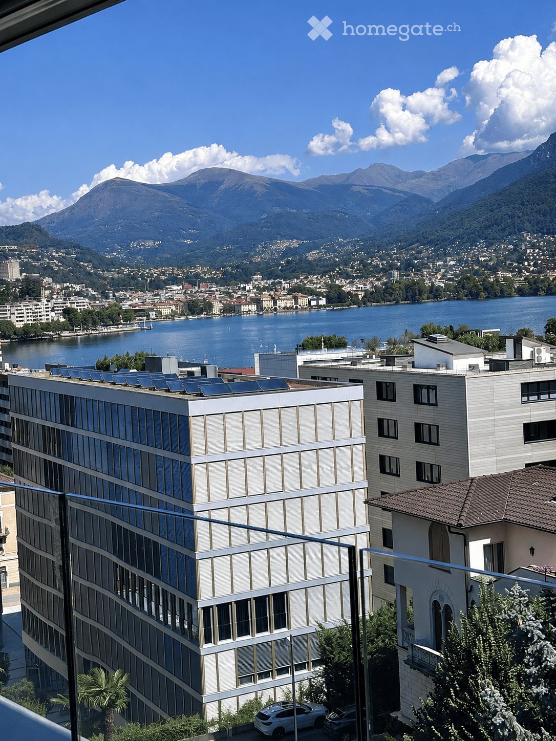 Balcony view overlooking a cityscape with a lake, mountains, and modern buildings.