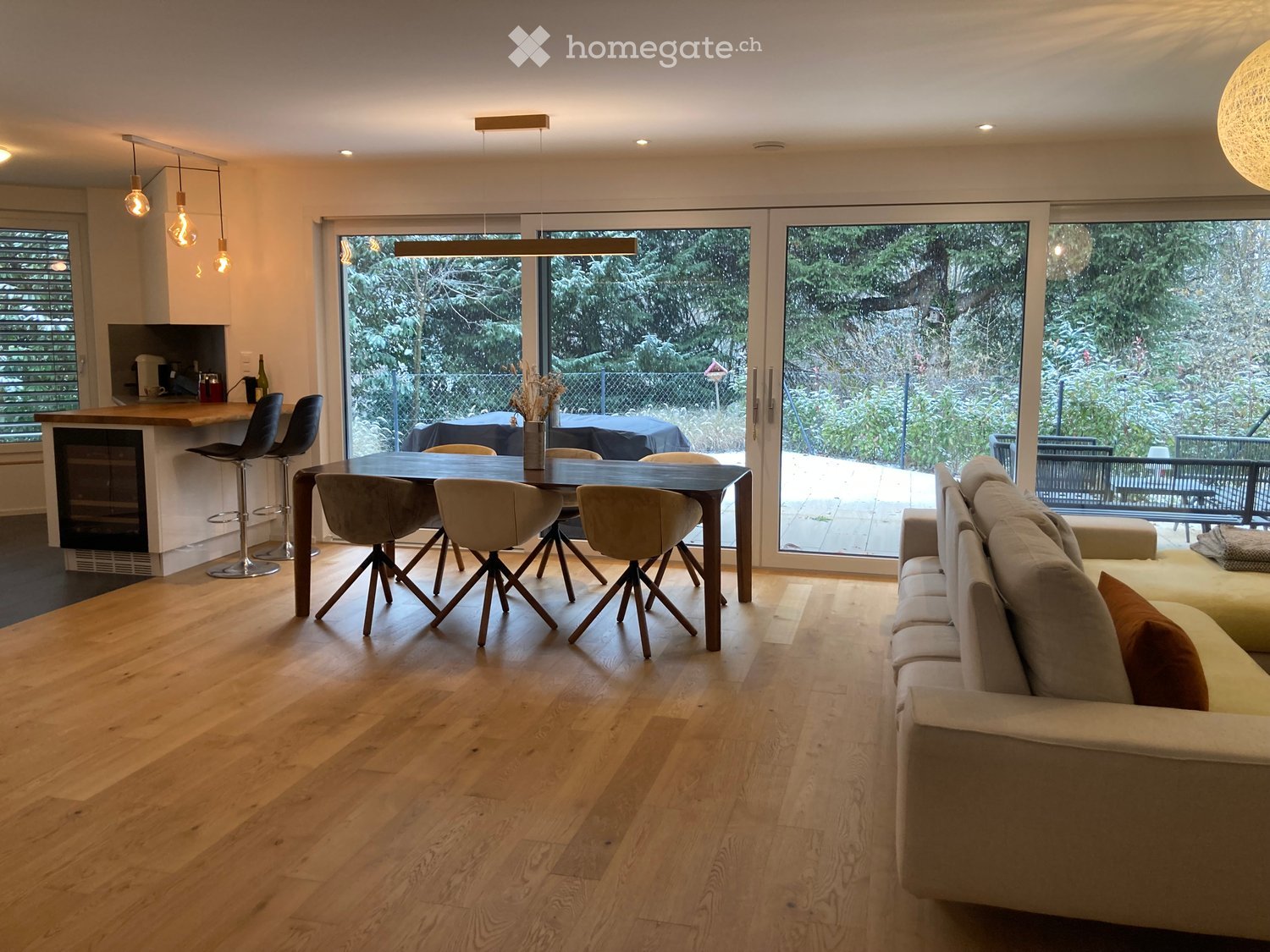 Open-plan living and dining area with light wooden flooring, a large sofa, a dining table with chairs, and a kitchen island with bar stools. Large windows offer a view of the garden.