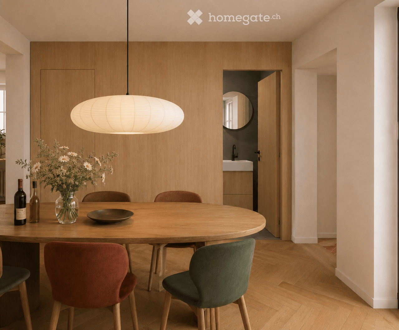 Modern dining area with a round wooden table, four chairs, a vase with flowers, two bottles, and a bowl on the table. A pendant light hangs above, and a doorway leads to a bathroom.