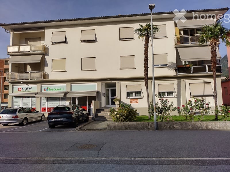 Three-story building with a modern facade, multiple windows with shutters, and a small entrance area. There are two cars parked in front, and the building has a small garden area with palm trees and shrubs.